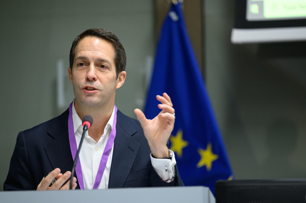 Man speaking into a conference microphone during a formal European Union meeting, gesturing with one hand, wearing a dark suit and purple lanyard, with a blurred EU flag and presentation screen in the background.